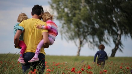 Father holding two children, as a third runs through flowers