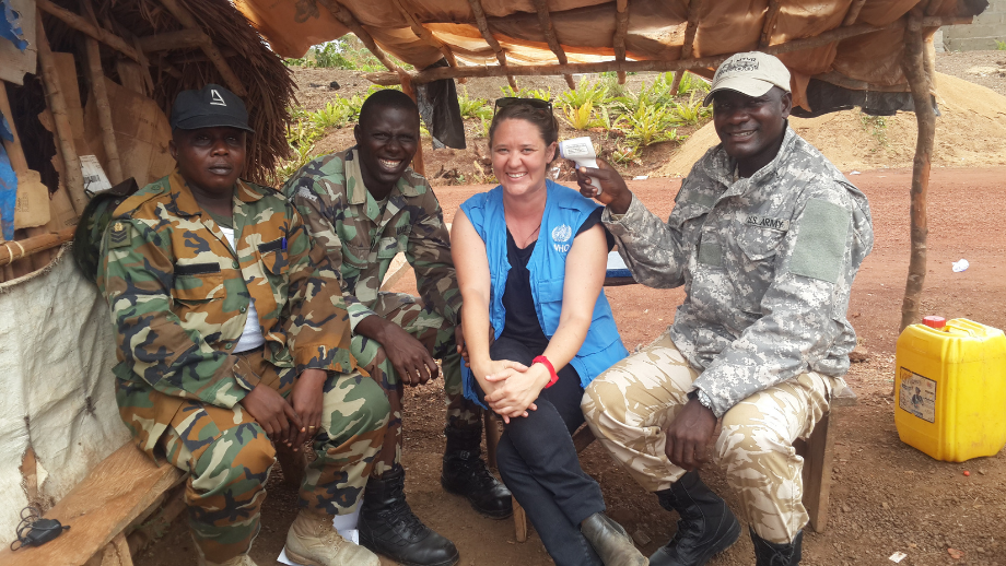 Three military personnel in camouflage uniforms and a woman in a blue vest are smiling and sitting under a makeshift shelter.