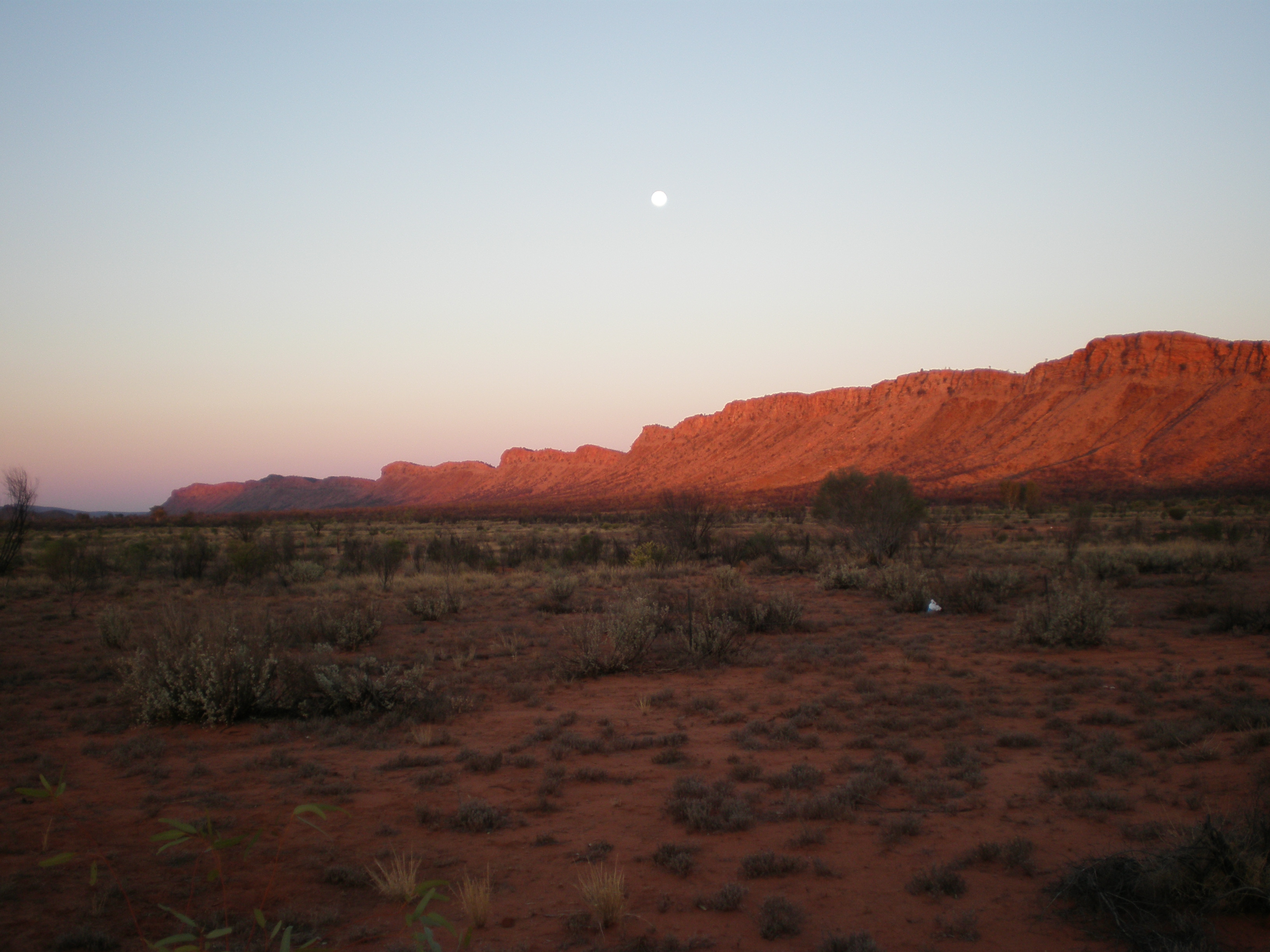 A view of a rugged, reddish mountain range glowing under the sunset in a vast, arid landscape with sparse vegetation under a clear sky.