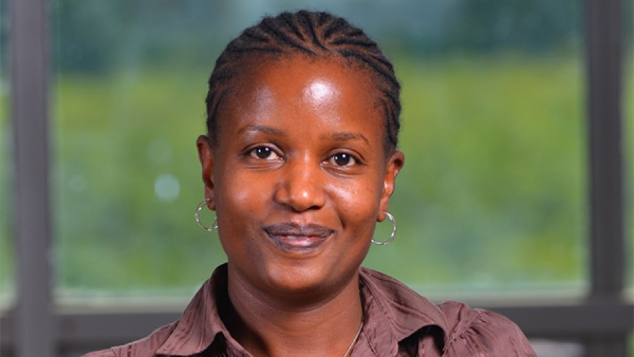 A woman with short braided hair, wearing a brown shirt and hoop earrings, smiling at the camera.