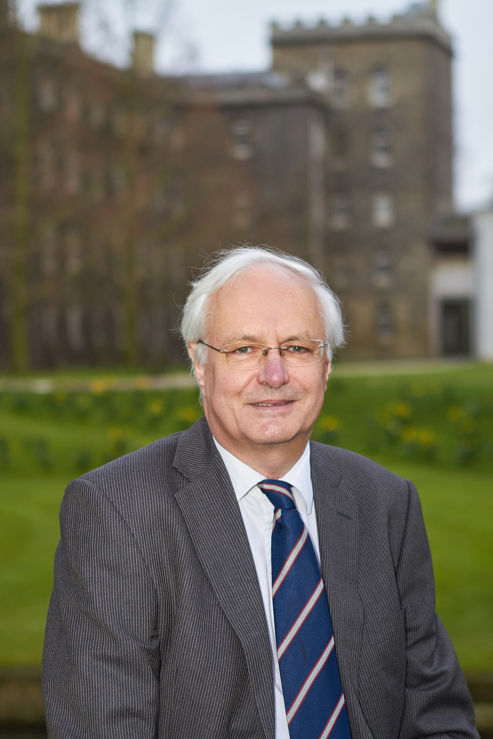 An older man in a suit and tie stands smiling in a park with historical buildings in the background.