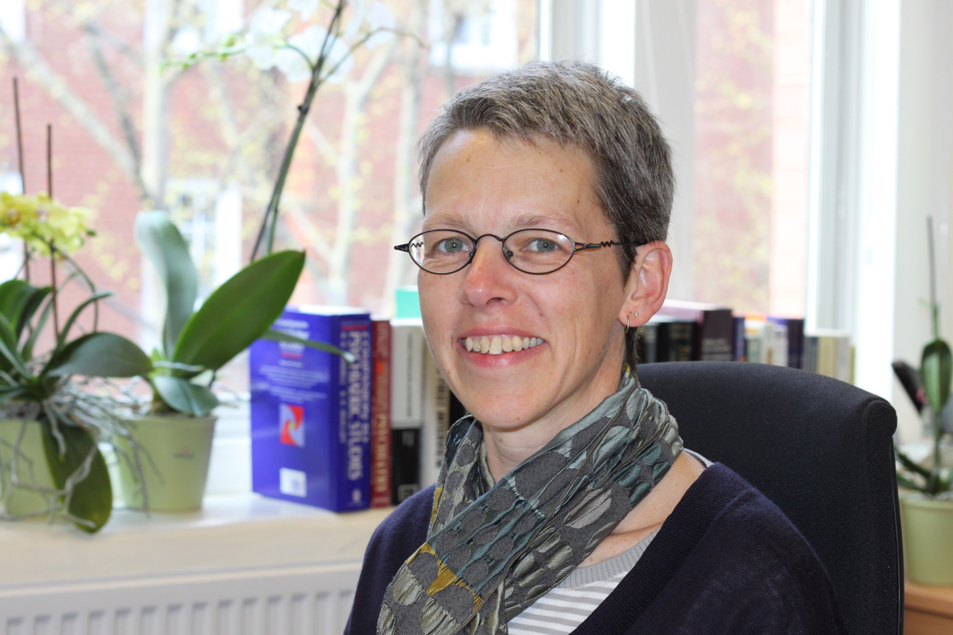 A woman with short gray hair and glasses, smiling in an office setting with books and potted plants in the background.