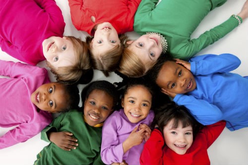 A group of seven diverse children wearing colorful shirts, lying in a circle with their heads together, smiling upwards.