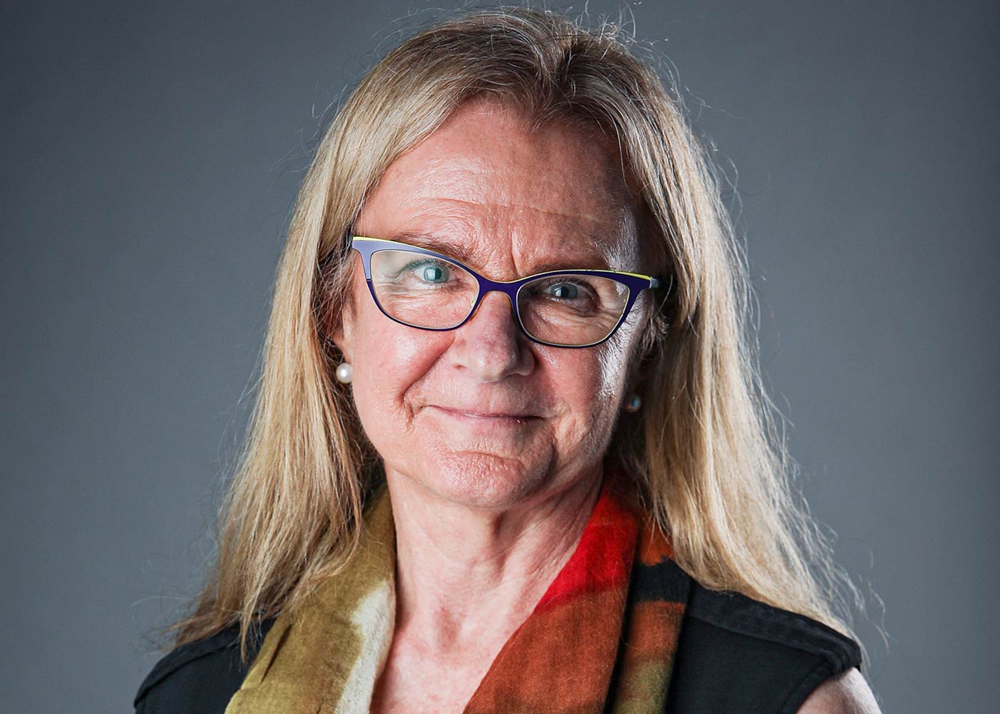 A portrait of an elderly woman with blond hair, wearing glasses, a black top, and a colorful scarf, smiling at the camera.