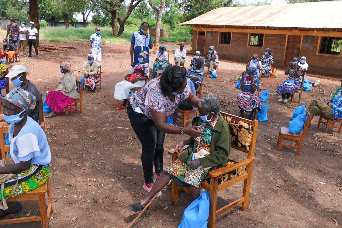 People at an outdoor community gathering in a rural setting with women and men seated on chairs, some wearing masks, as a woman assists an elderly lady.