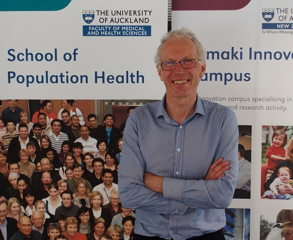 A man with glasses and a blue shirt smiling and standing with crossed arms in front of a banner that reads "School of Population Health" at the University of Auckland.