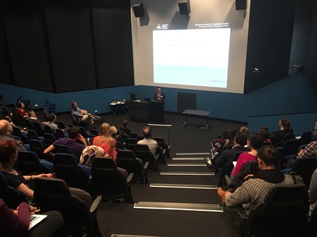 Audience attending a presentation in a modern lecture hall with a speaker at the podium.