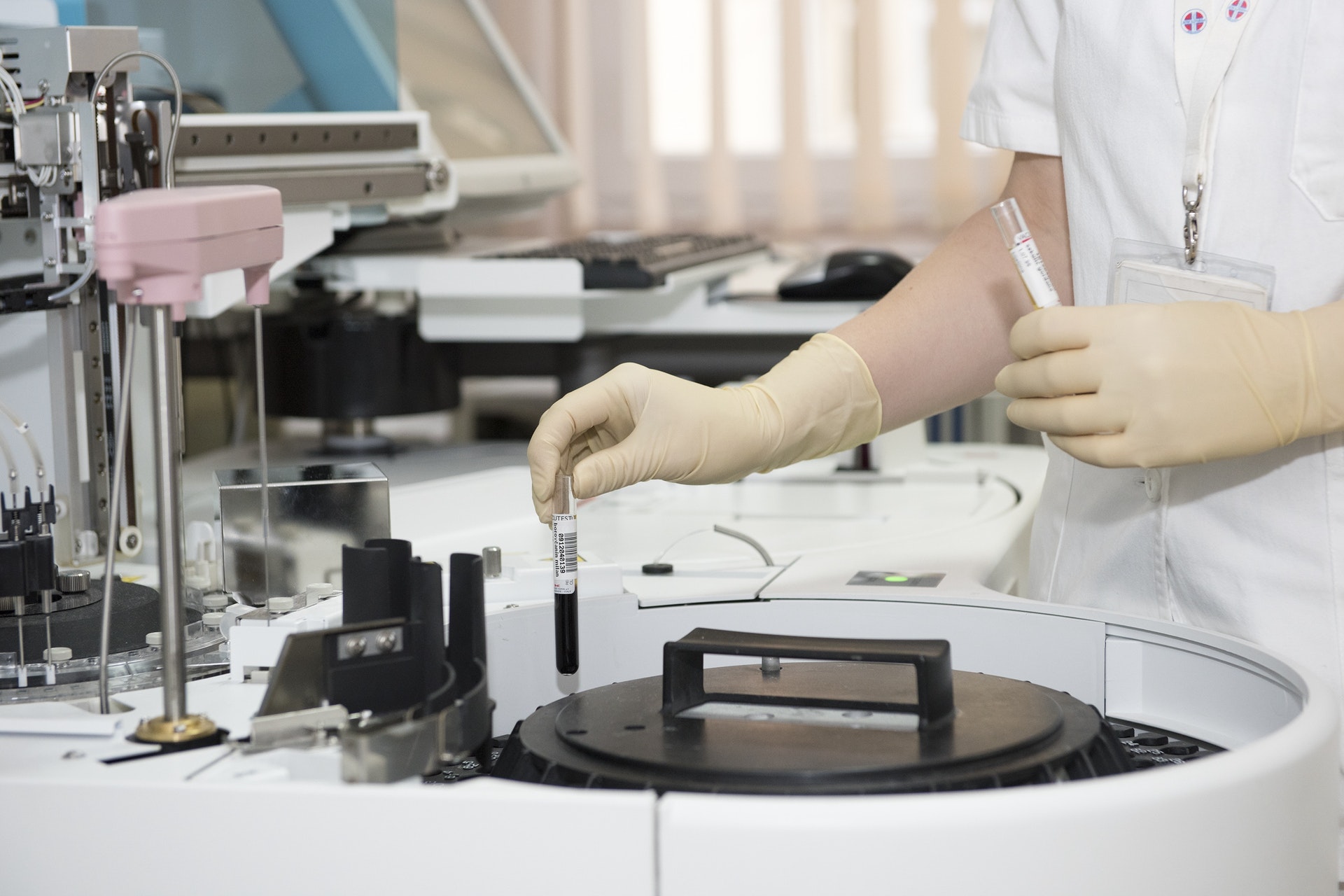 A technician in a laboratory wearing gloves places a test tube into an automated analyzer machine.