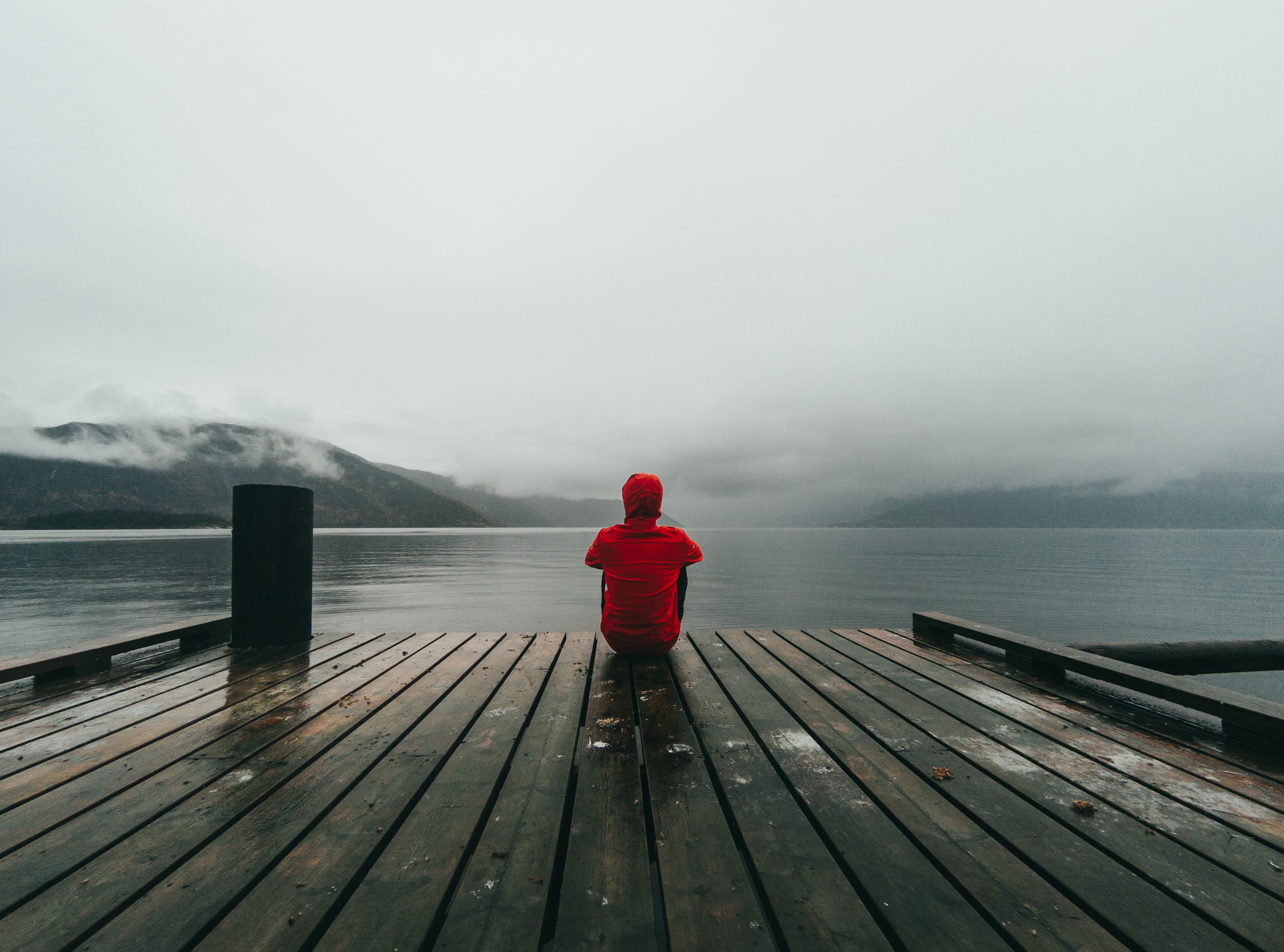 A person in a red hoodie sits at the end of a wooden dock, looking out at a foggy mountain lake.