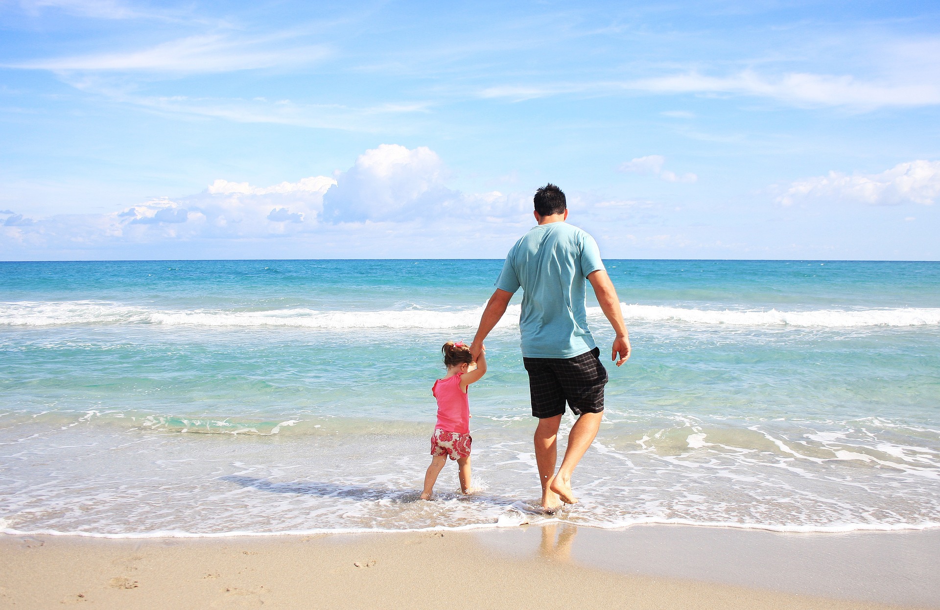 A man holding a young girl's hand on a sandy beach, both facing the sea under a clear blue sky.