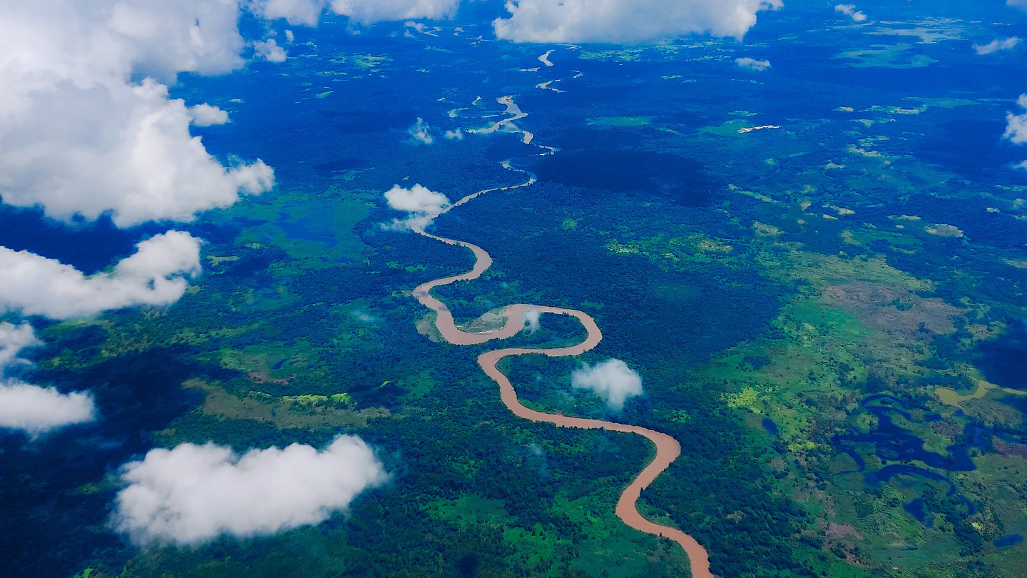 Aerial view of a meandering river winding through a dense green landscape with patches of clouds casting shadows on the ground.