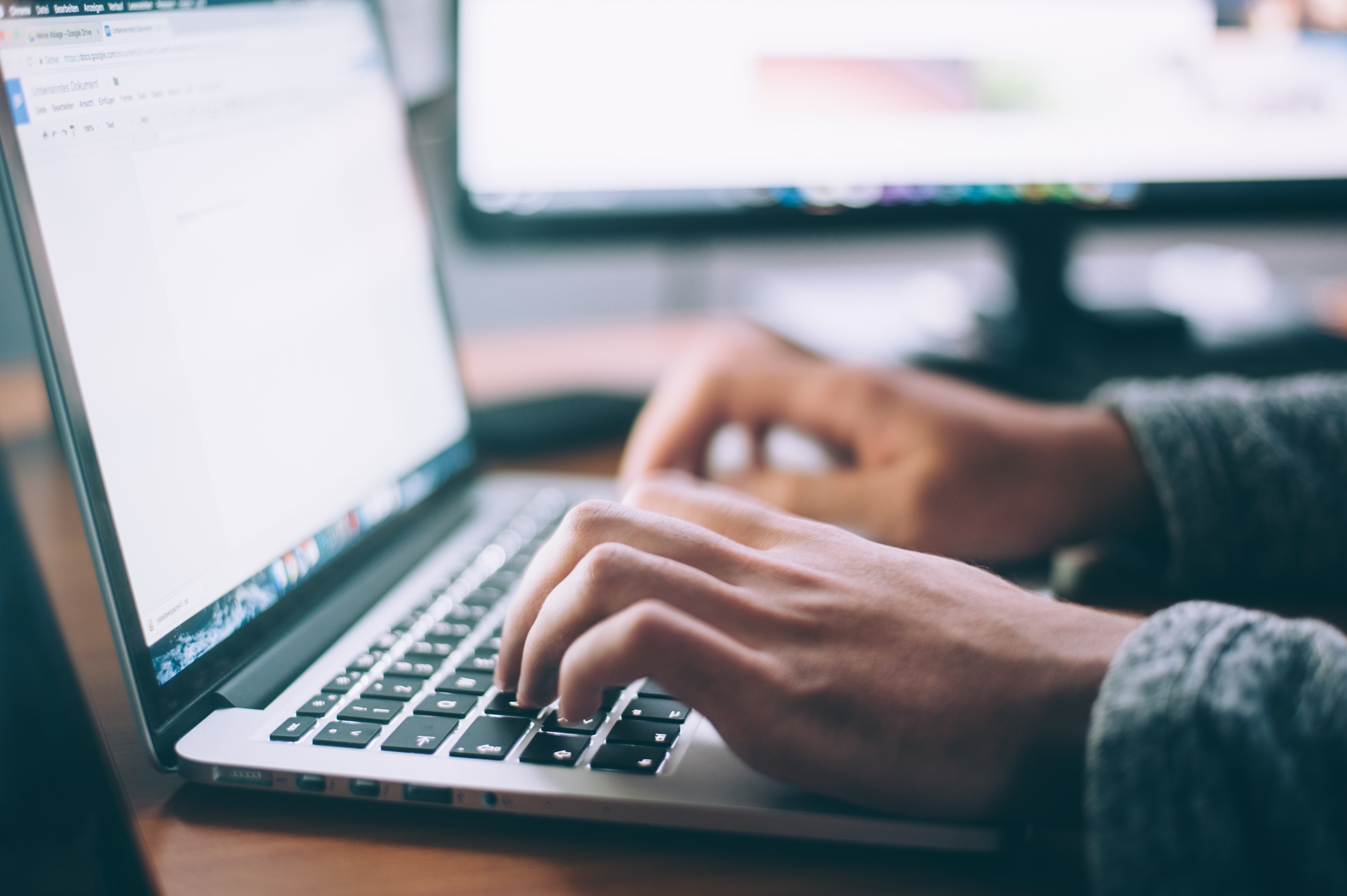 Person typing on a laptop in a softly lit room, with a blurry background featuring another computer screen.