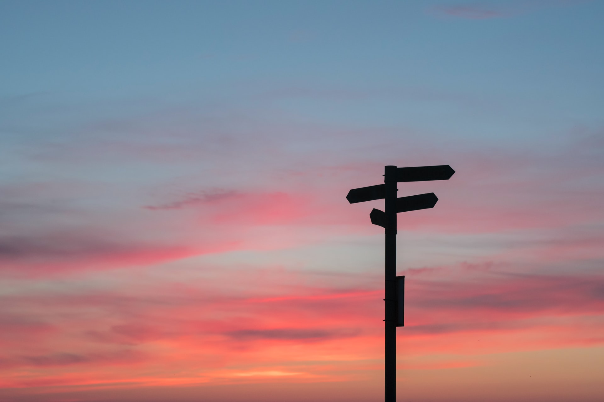 A silhouette of a directional signpost against a sunset sky with pink and blue hues.
