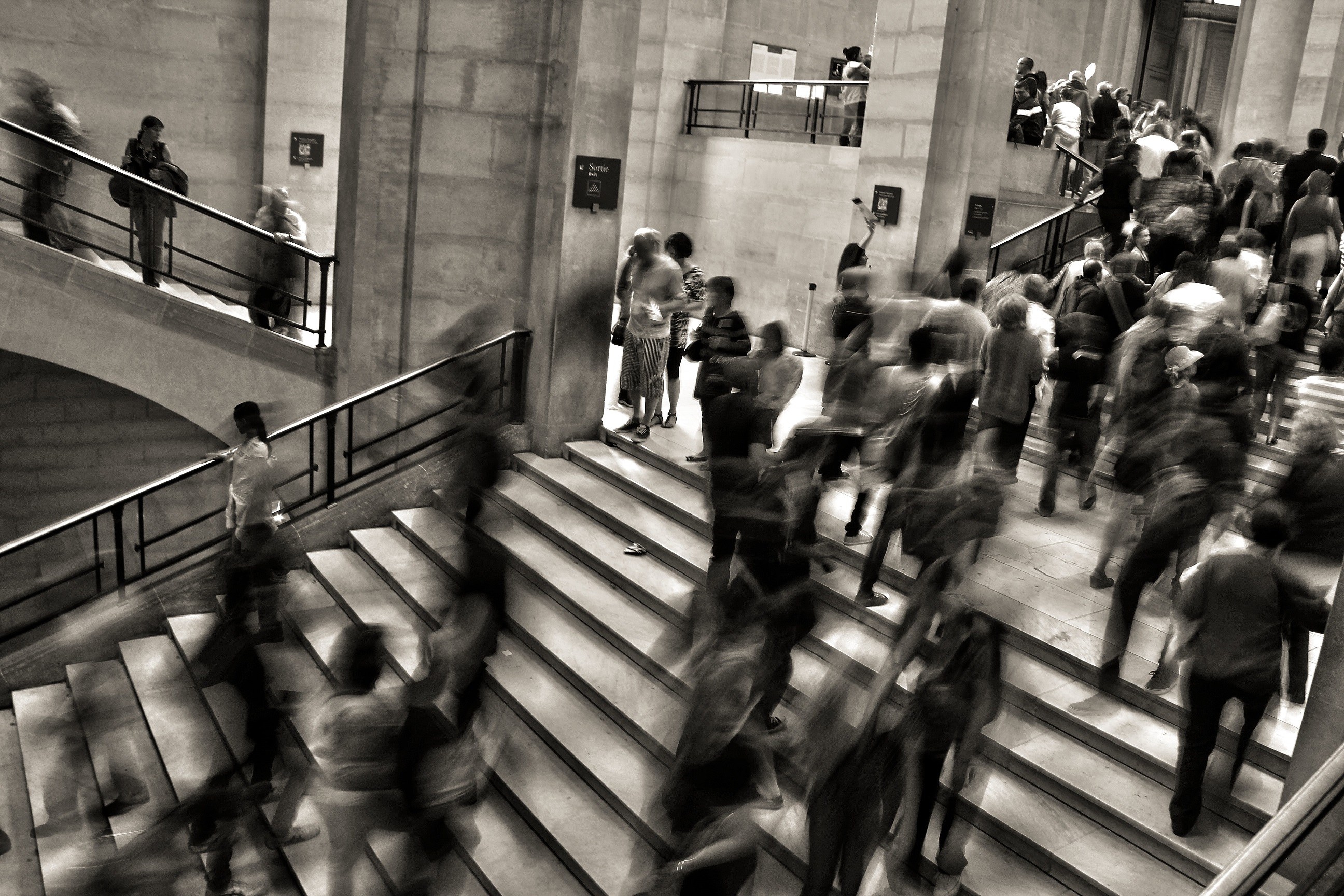 People moving rapidly up and down the grand staircase of a large building, captured in black and white.