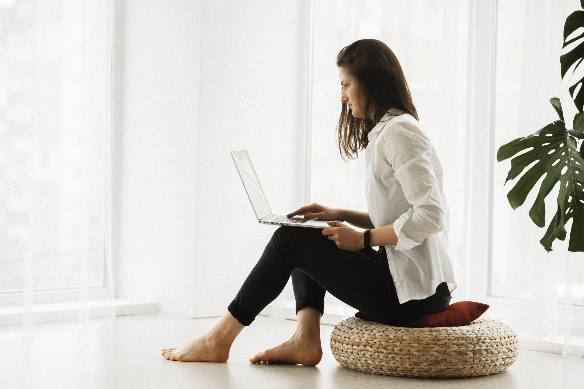A woman sits on a round woven ottoman using a laptop in a bright room with large windows and a green plant.