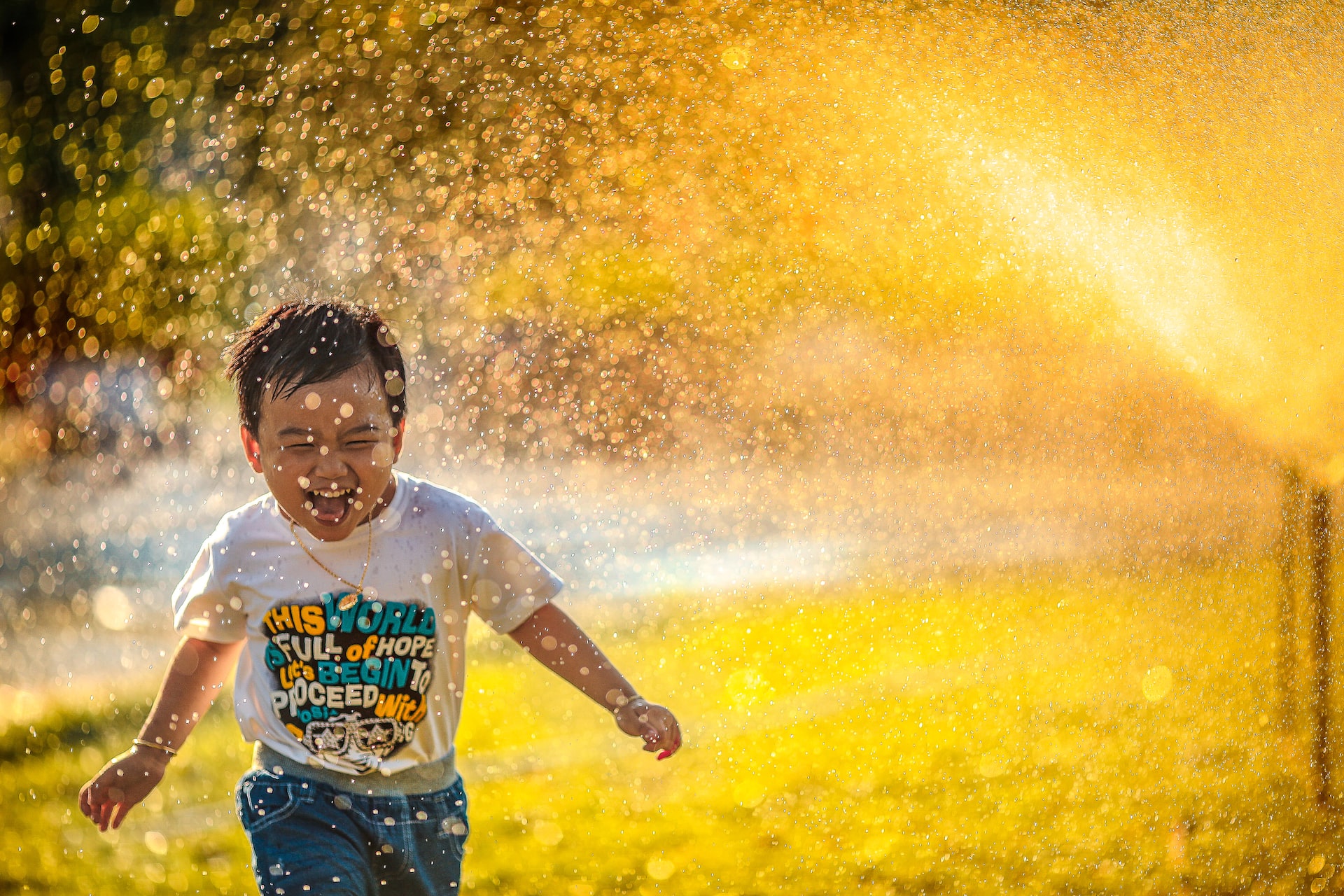 A young boy joyfully playing in sprinkling water with sunlight casting a golden glow around him.