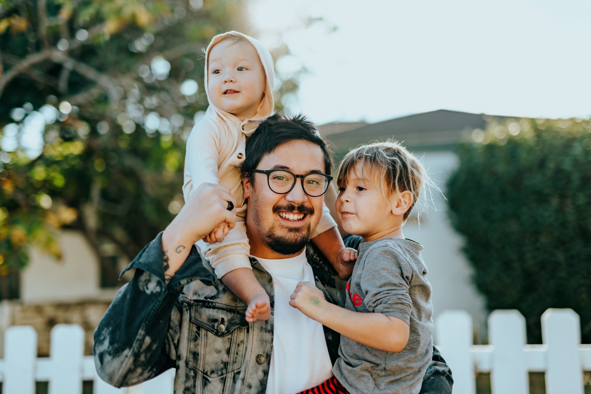 A joyful father carrying his two young children, one on his shoulders and the other by his side, outdoors near a white fence.