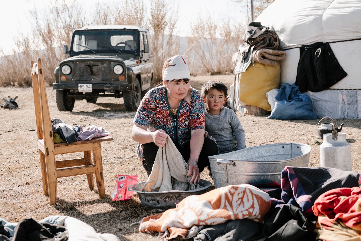 A woman washes laundry in a tub outdoors while a young child sits next to her, with a traditional tent and a rugged vehicle in the background.