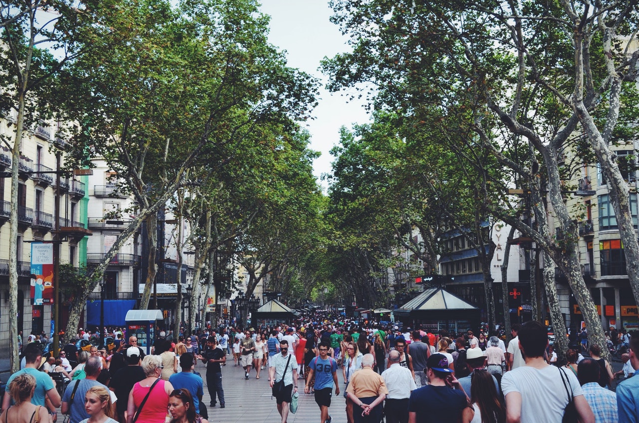 A bustling pedestrian street lined with trees and surrounded by traditional European buildings, filled with a diverse crowd of people.