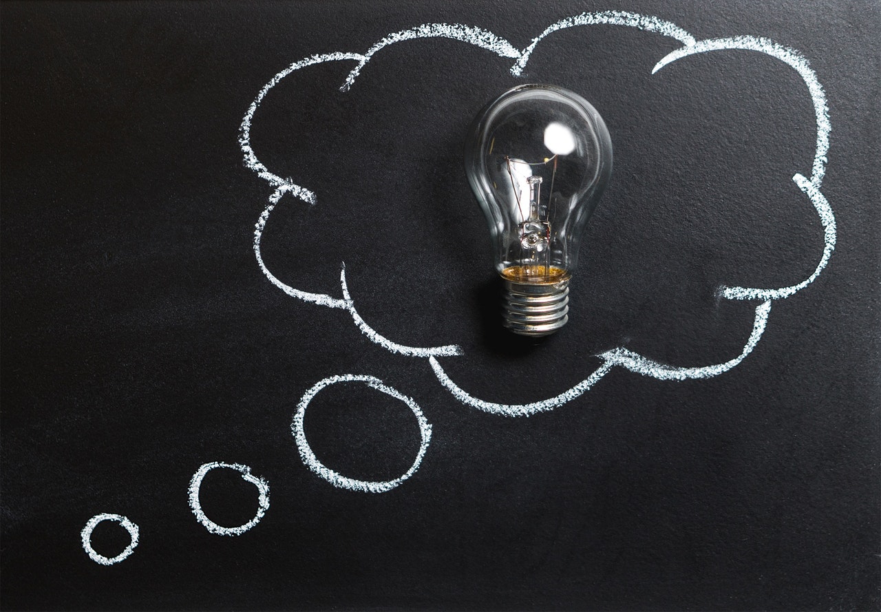 A clear lightbulb stands upright inside a white chalk thought bubble drawn on a blackboard.