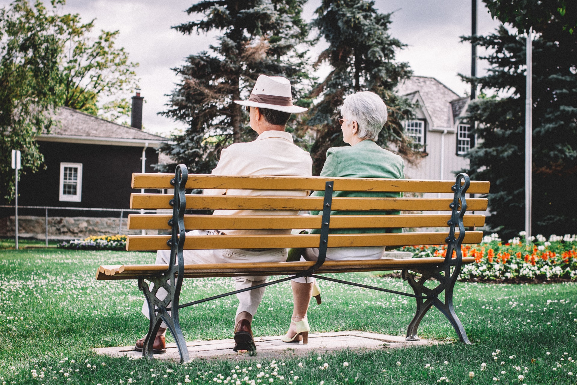 An elderly couple sits on a park bench, viewing a lush garden, with the woman wearing a green jacket and the man in a white hat.