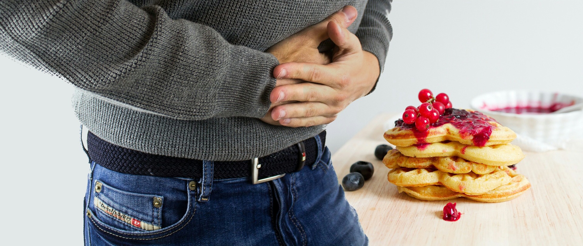 A person clutching their stomach with a plate of pancakes topped with berries in the background.