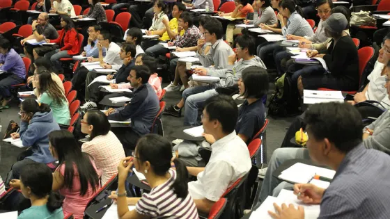 A diverse group of students sits in a university lecture hall, listening intently and taking notes.