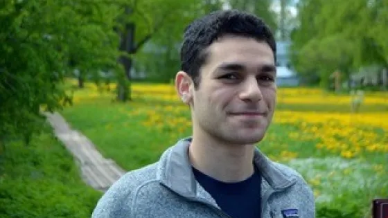 A young man smiling gently in a park with lush green trees and a field of yellow flowers in the background.