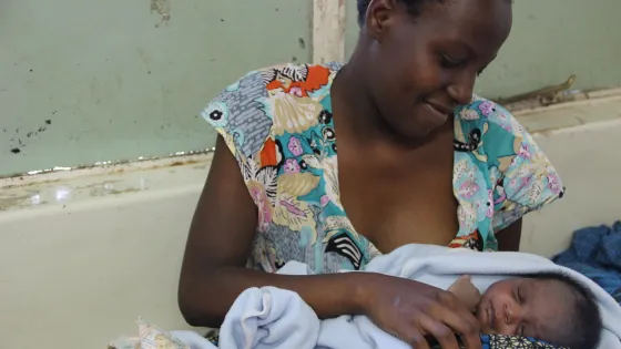 A woman smiles gently while holding a sleeping newborn baby wrapped in a light blue blanket.