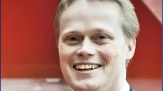 A portrait of a smiling man with blond hair, dressed in a dark suit, in front of a red background.