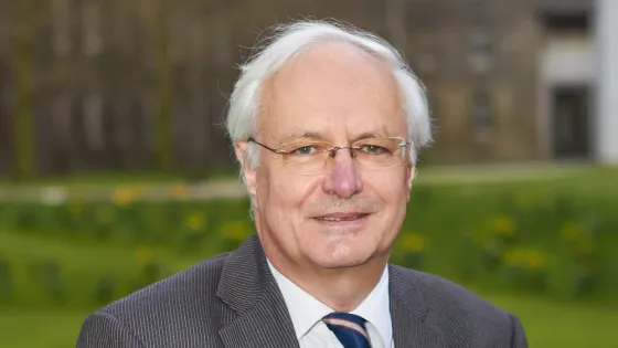 An older man in a suit and tie stands smiling in a park with historical buildings in the background.