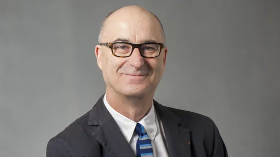 A smiling older man wearing glasses, a suit, and a striped blue tie poses against a gray background.