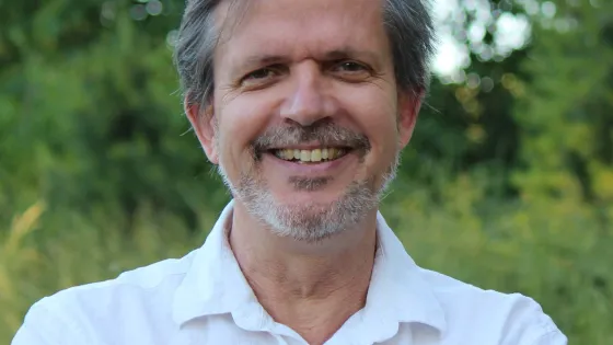 A smiling man with graying hair and a beard, wearing a white shirt, standing in front of a background of green foliage.