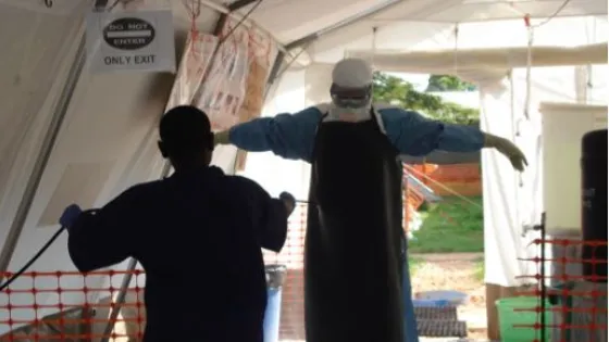 A healthcare worker in protective gear guides another person through a medical tent exit.