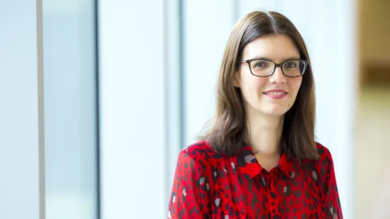 A woman in a red blouse with a black heart pattern stands smiling in a brightly lit office space.