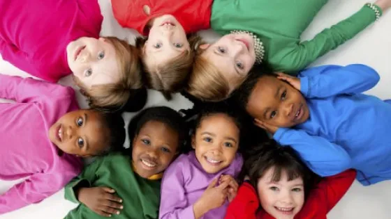 A group of seven diverse children wearing colorful shirts, lying in a circle with their heads together, smiling upwards.