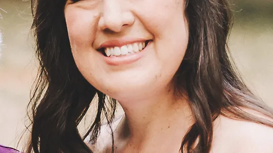 A smiling woman with side-swept bangs and long, curly hair wearing a white dress.
