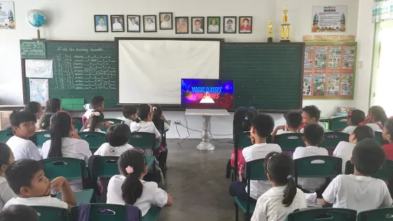 A classroom of students in uniforms paying attention to a video presentation on a television screen at the front of the room.