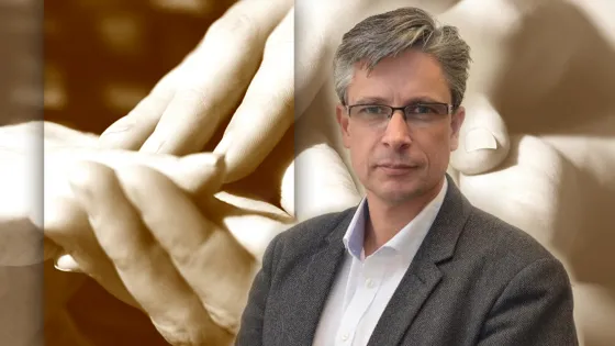 A man in glasses and a blazer stands in front of a blurred background, with a superimposed image of two hands almost touching, reminiscent of Michelangelo's "Creation of Adam."
