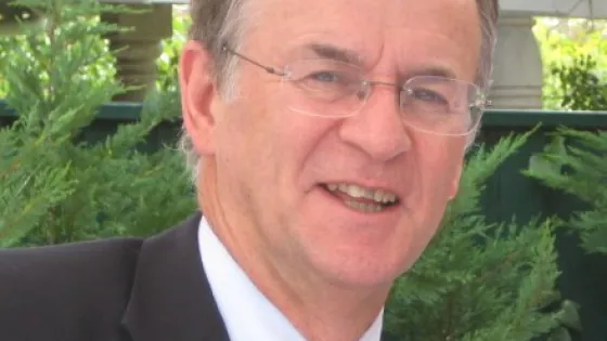 A smiling man in a suit and tie with greenery in the background.
