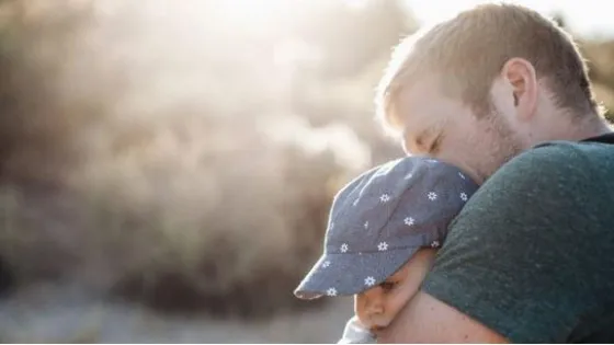 A man tenderly holds a sleeping child against his shoulder in a sunlit outdoor setting.