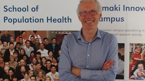A man with glasses and a blue shirt smiling and standing with crossed arms in front of a banner that reads "School of Population Health" at the University of Auckland.