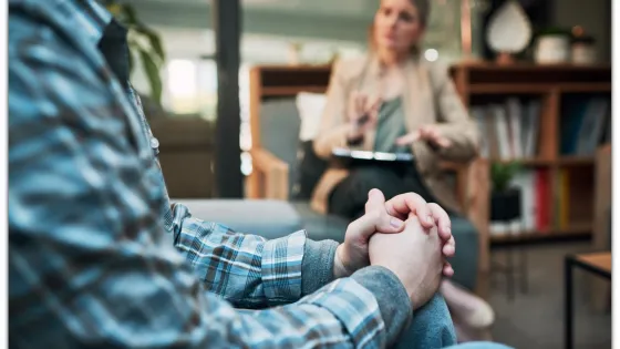 A man in a plaid shirt clasps his hands while talking to a woman in a beige blazer who is gesturing and holding a clipboard in a cozy office setting.