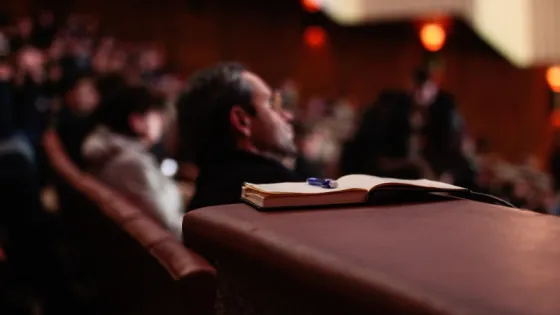 Audience members sitting in a dimly lit theater with a focus on an open notebook resting on an armrest.