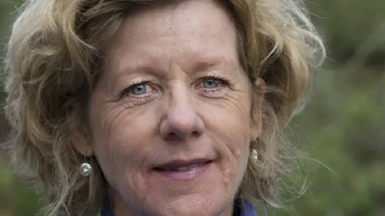 Portrait of a middle-aged woman with tousled blonde hair, wearing a blue turtleneck and small earrings, smiling at the camera.