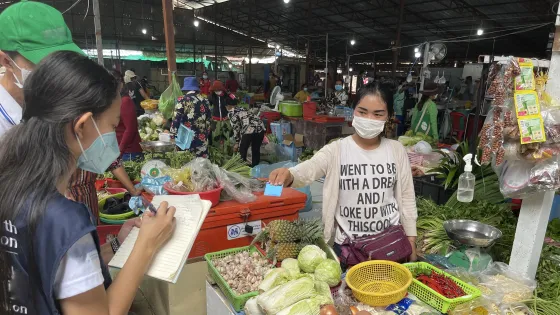 A bustling market scene with vendors and customers wearing masks, one vendor pointing at her sale scale, surrounded by fresh vegetables and goods.