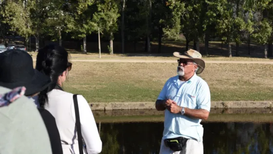 A man in a cowboy hat explains something to a group by the water's edge in a park.