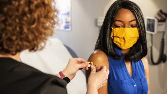 A healthcare worker applies a bandage to a woman's arm after a vaccination; both are wearing masks in a medical office.