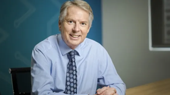 A smiling man in a blue shirt and patterned tie sitting at a desk in an office setting.