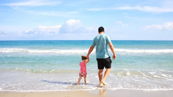 A man holding a young girl's hand on a sandy beach, both facing the sea under a clear blue sky.
